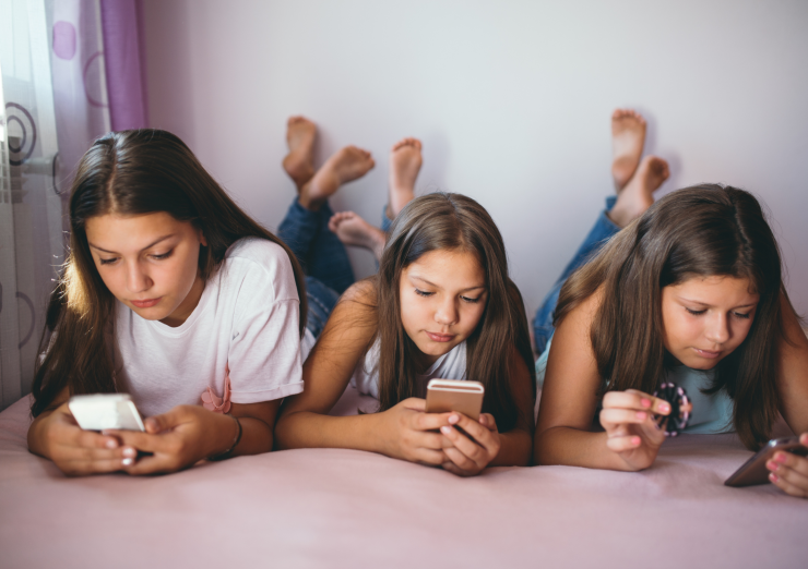 three teenage girls lying on a bed looking at their phones and not each other